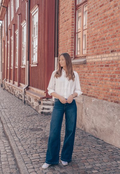 Camisa blanca de botones con mangas arremangadas, jeans de pierna ancha de mezclilla y zapatillas claras, de pie sobre una calle empedrada al lado de edificios de ladrillo rojo.