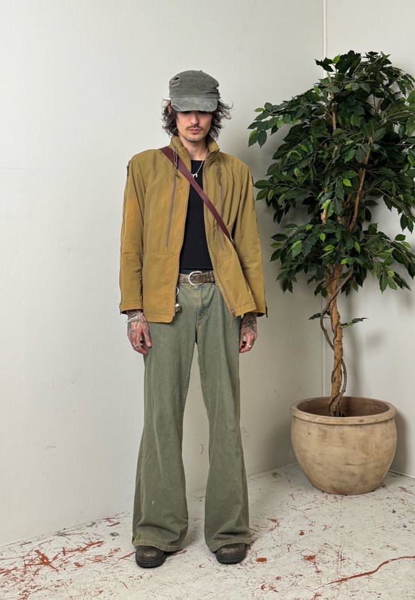Jeune homme portant une veste marron, un pantalon vert et une casquette grise, debout à côté d'une grande plante en pot contre un mur uni.