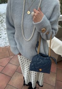 Personne portant un pull pelucheux bleu clair avec des boutons dorés, un collier de perles et une bague, une jupe blanche à pois, tenant un sac matelassé bleu marine avec une chaîne dorée.