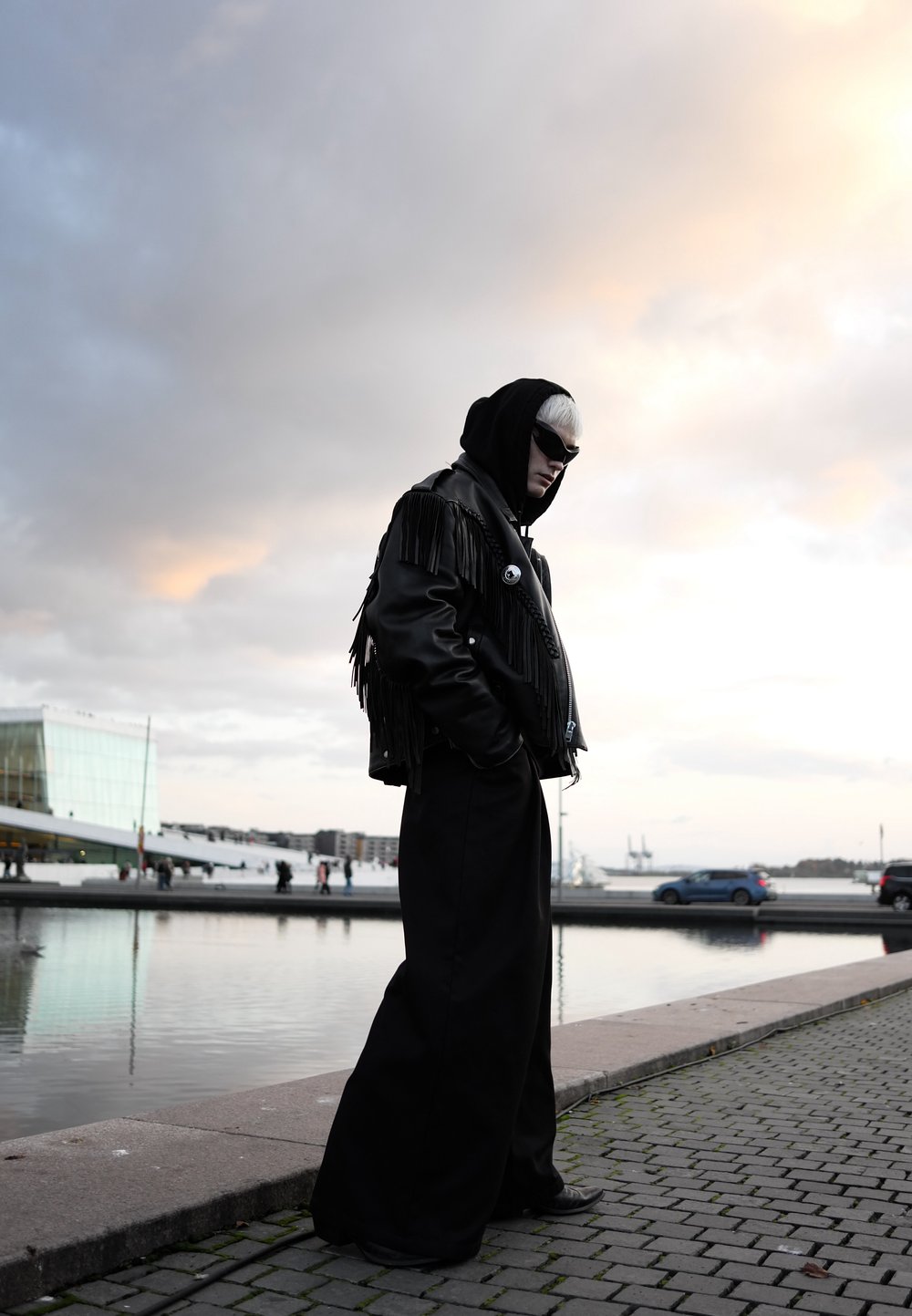 Person i svart fransad läderjacka, vida byxor, solglasögon och huva står vid vattnet med moderna byggnader och molnig himmel vid solnedgång.
