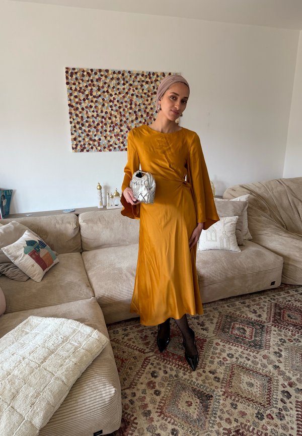 Femeie purtând o rochie galben muștar și un șal pe cap, ținând o geantă de mână argintie într-un living cu canapele bej și un covor cu model.