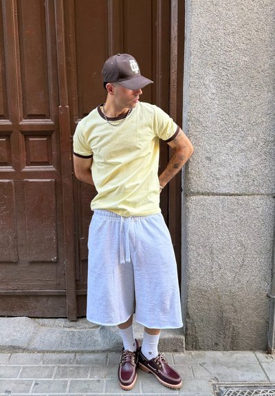 Hombre con camiseta amarilla, pantalones cortos blancos, gorra marrón, calcetines blancos y zapatos burdeos, de pie junto a una puerta marrón y una pared de piedra.