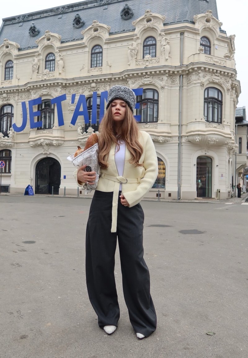 Giovane donna con cappello invernale e cappotto che tiene in mano pane e caffè, in piedi di fronte a un edificio ornato con un grande cartello "JE T'AIME".