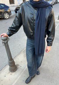 Blouson aviateur en cuir noir, écharpe bleu marine, jean en denim et bottines noires. La tenue présente une coupe décontractée et des textures simples.