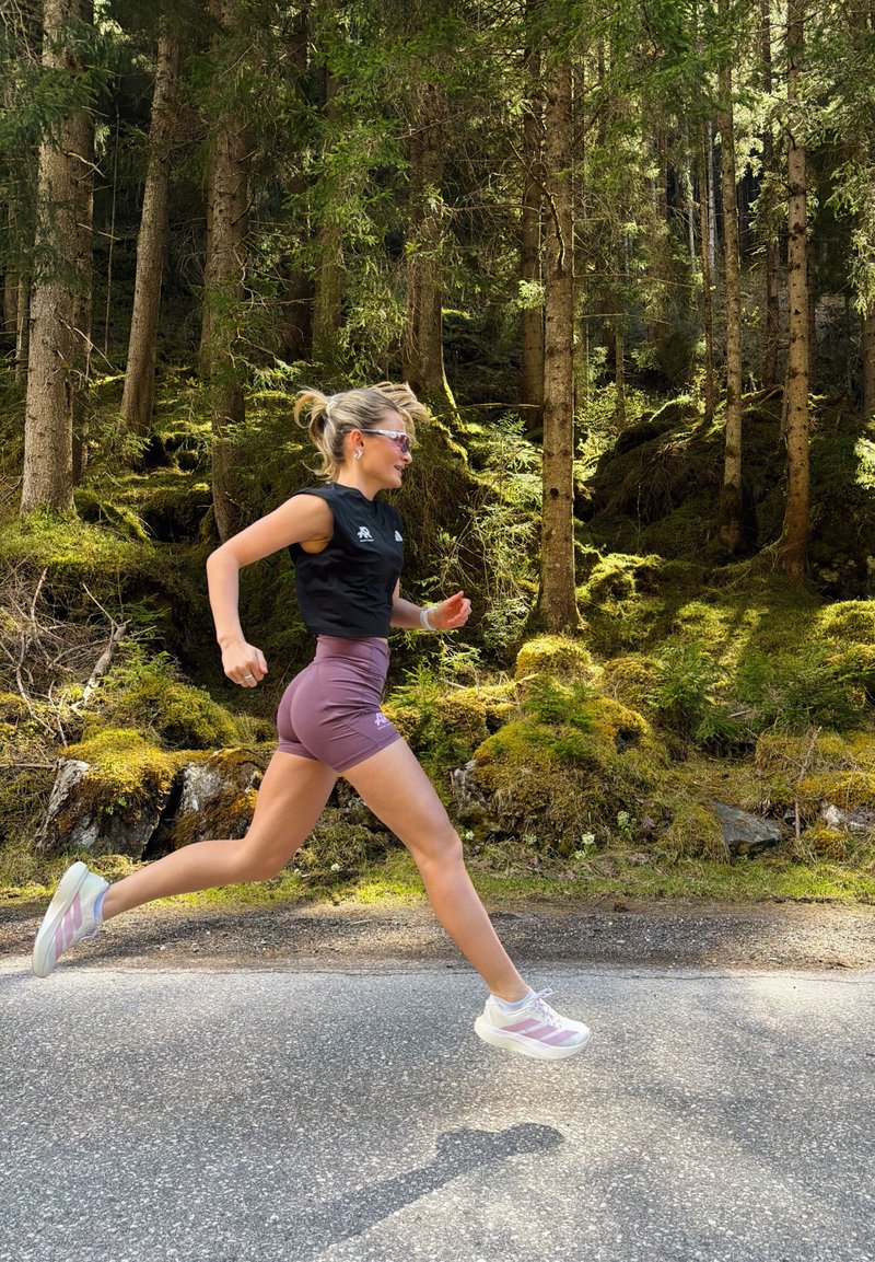Mujer con ropa deportiva corriendo a mitad de zancada por una carretera pavimentada junto a un bosque denso con musgo y luz del sol filtrándose a través de árboles altos.