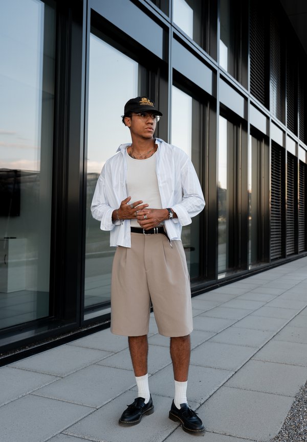 Jeune homme portant une chemise blanche, un short beige, une casquette noire, des lunettes et des chaussures noires, debout sur un trottoir à côté d'un bâtiment moderne en verre.