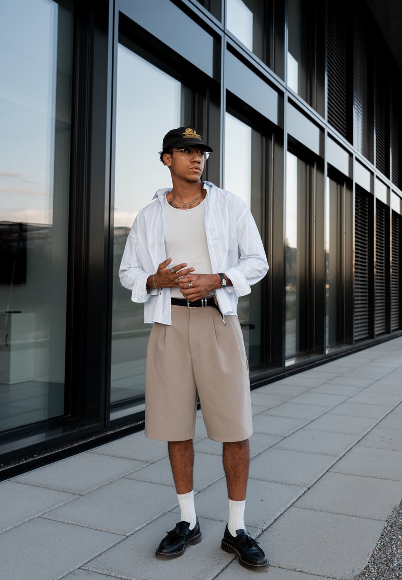 Joven con camisa blanca, pantalones cortos beige, gorra negra, gafas y zapatos negros, de pie en una acera junto a un edificio moderno de vidrio.