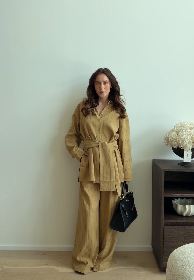 Femme aux longs cheveux bruns portant un costume beige à fines rayures, tenant un sac à main noir, debout contre un mur blanc près d'une armoire sombre avec des fleurs blanches.