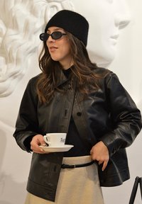 Veste en cuir noire à col, portée sur un haut noir. Tenant une tasse de café blanche sur une sous-tasse, portant des lunettes de soleil ovales foncées et un bonnet en tricot noir.