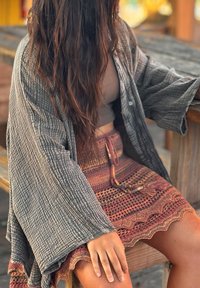 Γυναίκα με μακριά μαλλιά που φοράει γκρι υφασμένο ριχτάρι, τοπ σε χρώμα ταούπε και πολύχρωμη πλεκτή φούστα, καθισμένη σε ξύλινο παγκάκι σε εξωτερικό χώρο.