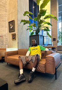 Zeleno-žlutý dres Adidas, maskáčové šortky, černé platformové boty; sedící na hnědé kožené pohovce v krytém prostoru s rostlinami.