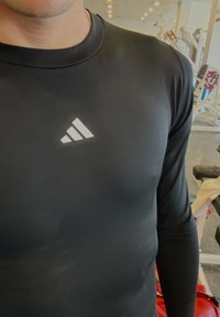 Fekete sportos hosszú ujjú felső, testhezálló szabással. Az elején fehér Adidas logó látható. Az anyag sima és rugalmas hatású.