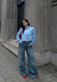 Chemise à manches courtes bleu clair, ornée de nœuds sur les manches, associée à un jean bleu évasé et des escarpins rouges vif à bout pointu. Fond en pierre.