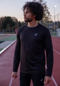 Mann mit lockigem Haar, der ein schwarzes Asics Langarmshirt und Leggings trägt und bei Dämmerung auf einer Außenlaufbahn steht.