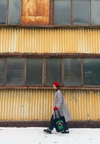 Femme en manteau gris et béret rouge marchant sur la neige à côté d'un mur en métal ondulé jaune, portant un sac fourre-tout noir avec un imprimé coloré.