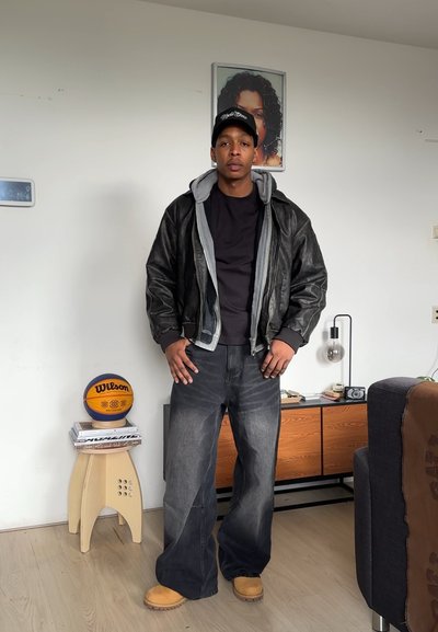 Chaqueta de cuero negra con capucha gris, camiseta negra, jeans oscuros sueltos y botas marrones. Un balón de baloncesto Wilson reposa sobre una mesa pequeña cercana.