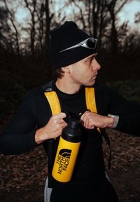Fekete hosszú ujjú ing és sárga hátizsákpántok; egy sárga, hőszigetelt vizespalackot tart fekete logóval; erdős háttér.