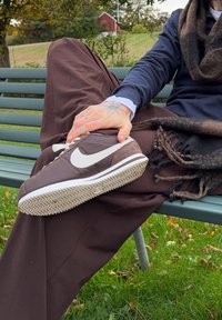 Barna velúr és bőr sneaker fehér Swoosh-sal, lapos talppal és halszálka mintával. Zöld parki háttér előtt.