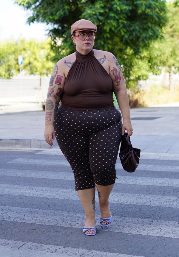 Persona con tatuajes que lleva un top marrón sin tirantes, pantalones de lunares, gorra y sandalias caminando por una calle sosteniendo una bolsa pequeña.