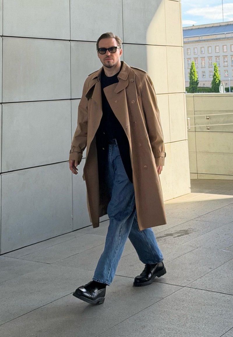 Homme portant des lunettes de soleil, un trench-coat marron, des chaussures noires et un jean bleu, marchant dehors près d’un mur de bâtiment moderne.