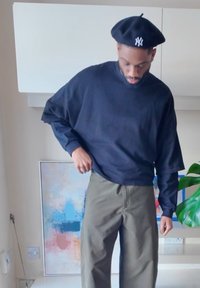 Čierna košeľa s dlhým rukávom a voľným strihom, doplnená olivovozelenými nohavicami. Čierny baret s bielym logom zvýrazňuje ležérny vzhľad.