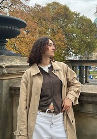 Μπεζ παλτό σε στιλ trench, επικαλυμμένο με καφέ κάρντιγκαν, συνδυασμένο με λευκά παντελόνια. Το σύνολο διαθέτει υφασμάτινη υφή και διακριτές χρυσές λεπτομέρειες στους κουμπιά.