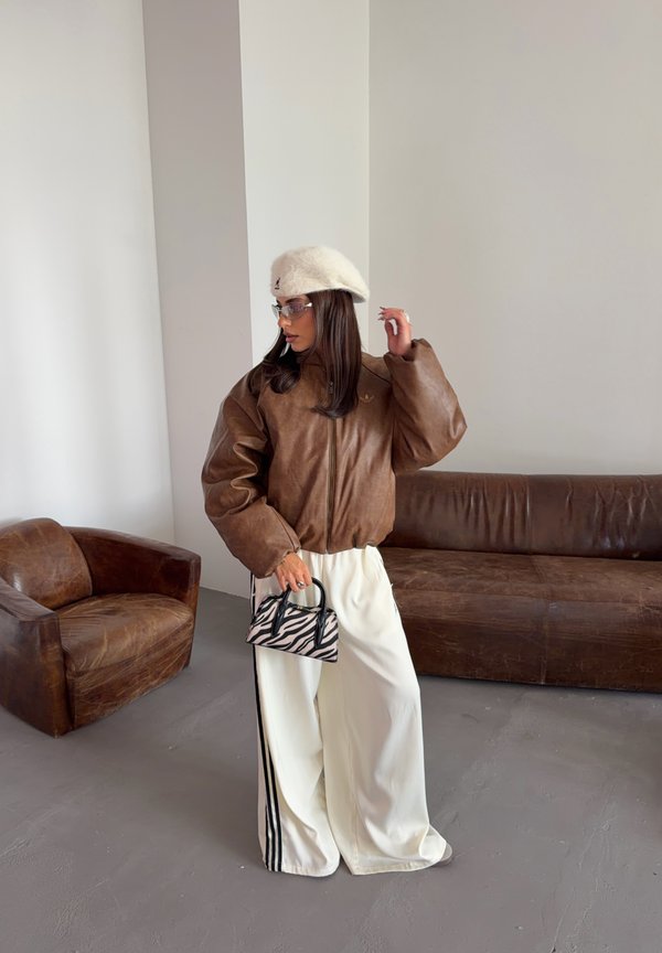 Mujer con chaqueta de cuero marrón, pantalones anchos blancos, boina blanca y gafas sosteniendo un bolso con rayas de cebra en una habitación con muebles de cuero marrón.