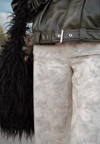 Tmavě zelená kožená bunda s páskem v pase, kombinovaná se světle šedými kalhotami s jemným květinovým vzorem a dlouhou černou chlupatou kabelkou.