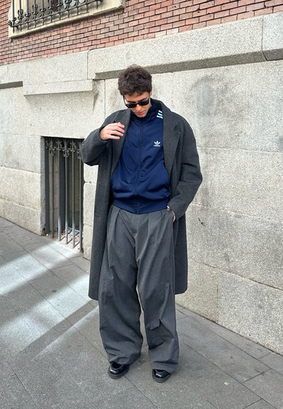 Chaqueta con cremallera Adidas en azul marino, abrigo gris oversize y pantalones grises anchos con pliegues. Zapatos negros, gafas de sol y fondo de pared de piedra texturizada.