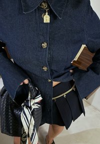Jeansjacke mit großem Kragen und dunklen Knöpfen, kombiniert mit einem geplisséten schwarzen Rock, einer gewebten schwarzen Tasche und einem Schal mit Blumenmuster.