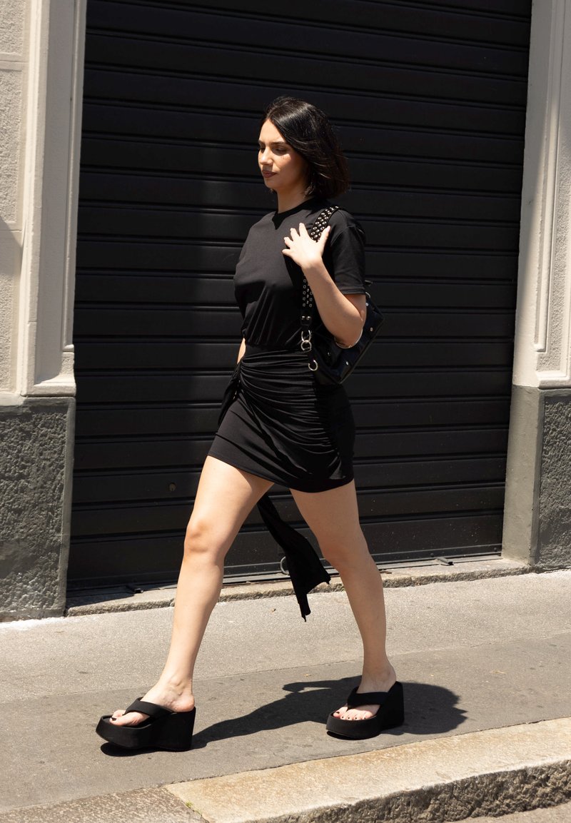 Μαύρο κοντό φόρεμα με εφαρμοστή γραμμή, υφή υφάσματος και λεπτομέρεια με κόμπο στο πλάι. Σανδάλια wedge σε μαύρο χρώμα με φαρδιές λουρίδες. Μαύρη τσάντα με τρουκς.