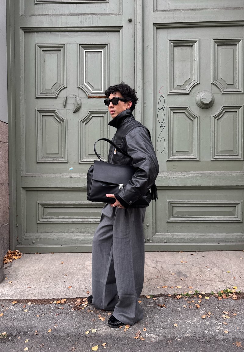 Black leather jacket with high collar, gray wide-leg trousers, black sunglasses, and a black bag with a handle and fringe detail, set against green doors.
