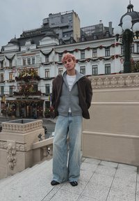 Jeune homme aux cheveux teints en rose se tenant sur des marches, portant un sweat à capuche gris, une veste marron et un jean large, avec un bâtiment historique décoré en arrière-plan.