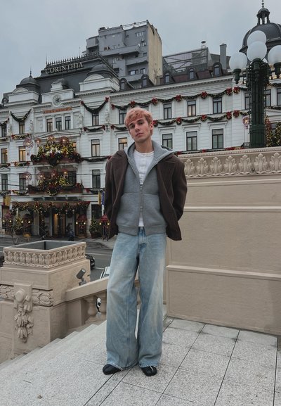 Joven con el cabello teñido de rosa de pie en unas escaleras, vistiendo una sudadera gris con capucha, chaqueta marrón y vaqueros de pierna ancha, con un edificio histórico decorado de fondo.