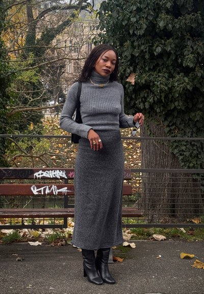 Suéter gris de cuello alto con canalé, falda larga ajustada a juego y botas negras de tacón hasta la rodilla, complementados con gafas de sol y una cadena dorada.
