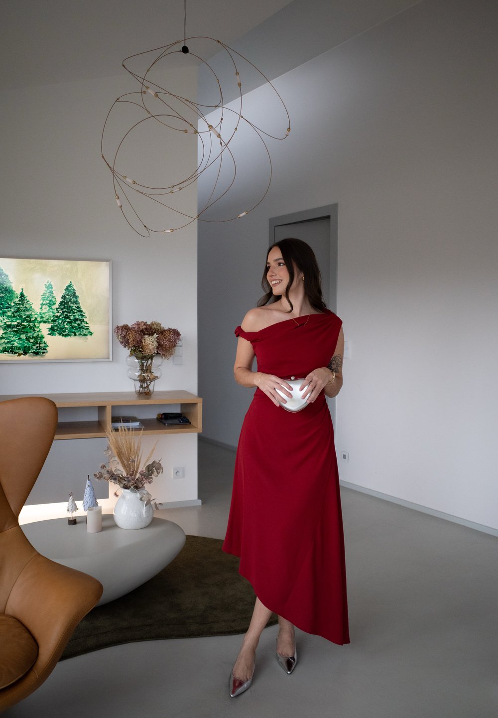 Κόκκινο φορεμα με ανοιχτούς ώμους και ριχτό σχέδιο, μαλακό ύφασμα. Ασημί ψηλές γόβες. Μοντέρνος εσωτερικός χώρος με φωτιστικό κρεμαστό και διακοσμητικά στοιχεία.