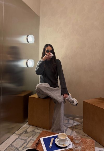 Mujer con gafas de sol y ropa casual sentada con las piernas cruzadas sobre una caja, bebiendo de una taza pequeña en una esquina moderna de un café.