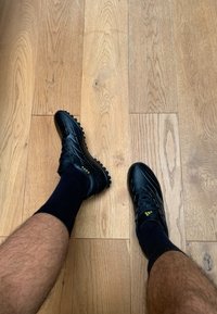 Zwarte voetbalschoenen met een gestructureerde oppervlak, met gele accenten. Draag met donkere sokken, geplaatst op een houten vloer.