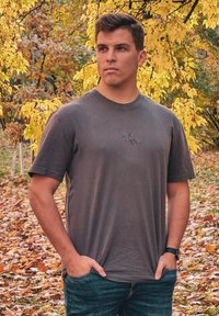 Graues Baumwoll-T-Shirt mit kurzen Ärmeln und Rundhalsausschnitt, präsentiert mit einem dezenten Logo in der Mitte. Getragen mit blauen Jeans in einer Außenumgebung.