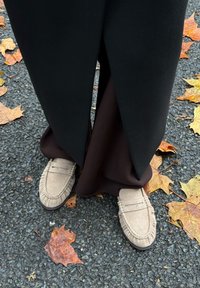 Bézs színű velúr mokaszinok varrással és pénznyílásos dizájnnal, barna szövetnadrággal párosítva, egy texturált sötét felületen, levelekkel.