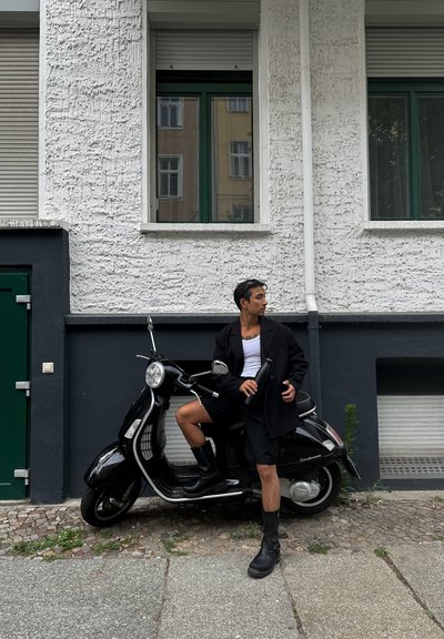 Scooter vintage negro con acentos de cromo, aparcado sobre adoquines. Persona con abrigo negro, camiseta blanca y botas negras, sosteniendo una botella.