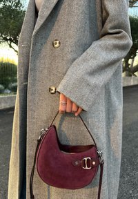 Sac à main en daim bordeaux à la forme courbée, doté d'une bandoulière en cuir marron et de ferrures argentées, tenu dans une main à côté d'un manteau gris à motif chevrons.