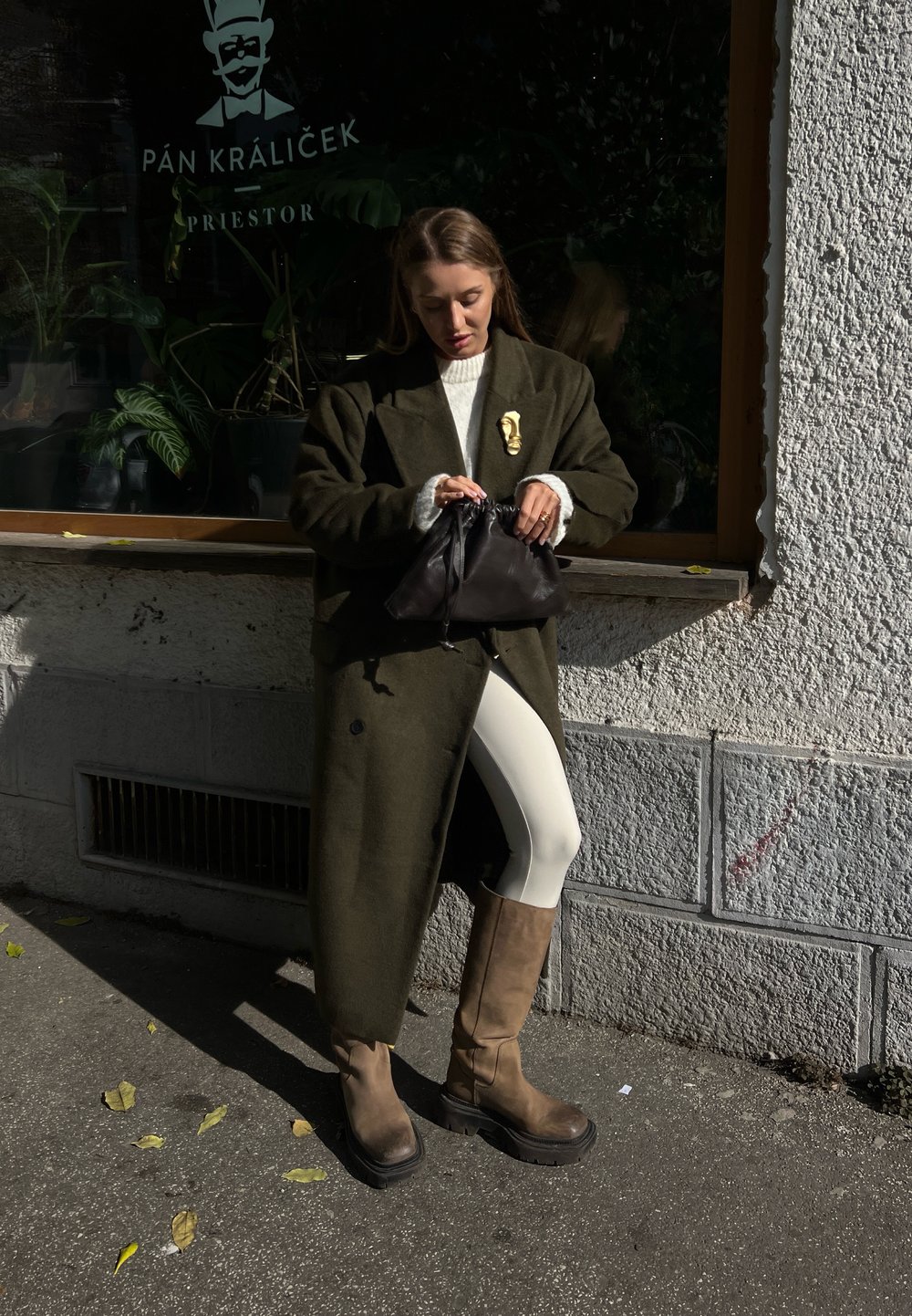 Πράσινο ελαιόλαδο μάλλινο παλτό, κρεμ πουλόβερ, λευκά κολάν και καφέ μπότες ως το γόνατο. Μαύρη τσάντα με μαζεμένο επάνω μέρος. Αστική τοποθεσία με παράθυρο.