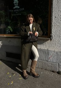 Πράσινο ελαιόλαδο μάλλινο παλτό, κρεμ πουλόβερ, λευκά κολάν και καφέ μπότες ως το γόνατο. Μαύρη τσάντα με μαζεμένο επάνω μέρος. Αστική τοποθεσία με παράθυρο.