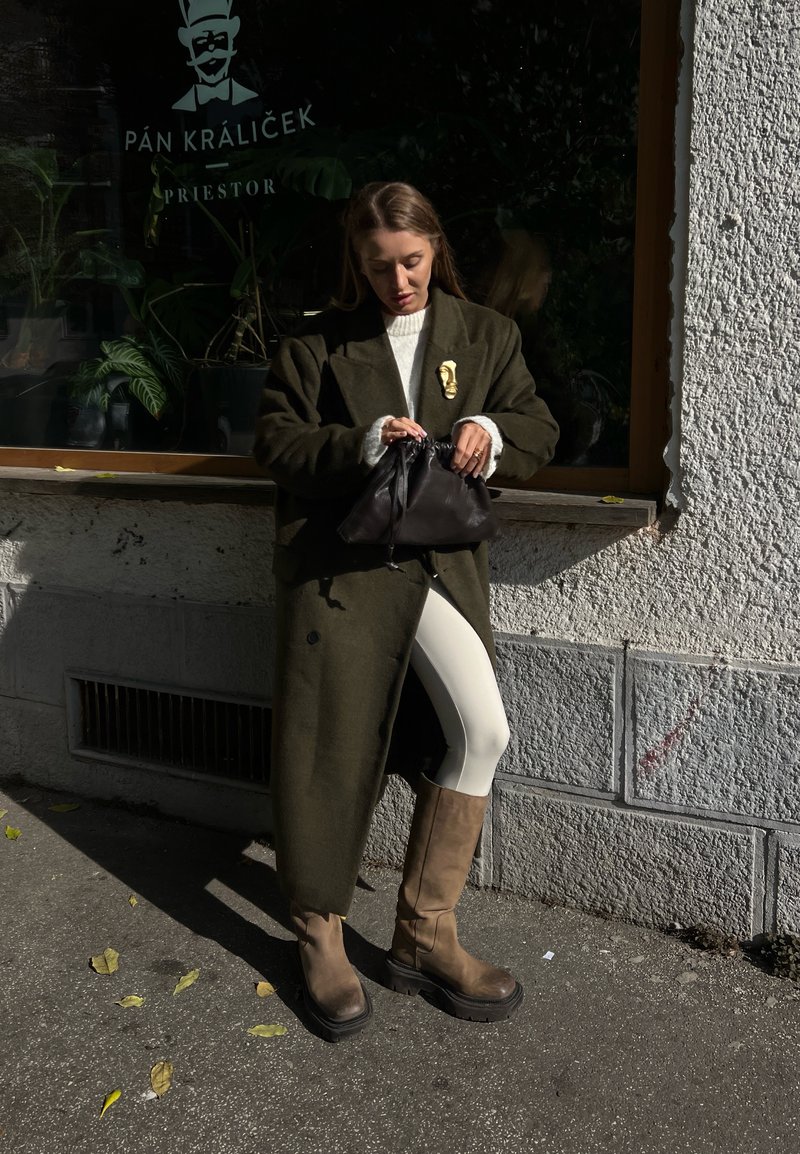 Πράσινο ελαιόλαδο μάλλινο παλτό, κρεμ πουλόβερ, λευκά κολάν και καφέ μπότες ως το γόνατο. Μαύρη τσάντα με μαζεμένο επάνω μέρος. Αστική τοποθεσία με παράθυρο.
