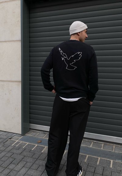 Sudadera negra con un contorno blanco de un pájaro en la parte posterior, combinada con pantalones negros y un gorro claro, en un entorno urbano.