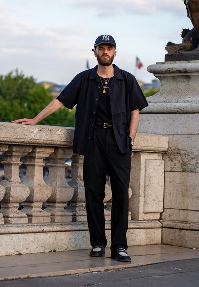 Camisa negra de manga corta, pantalones negros y zapatillas. Los accesorios incluyen collares y una gorra oscura con un logo blanco. Ambiente al aire libre.
