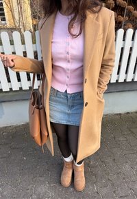 Manteau en laine de chameau, gilet rose à boutons, mini-jupe en denim, collants noirs, bottes UGG couleur beige et sac fourre-tout en cuir marron.