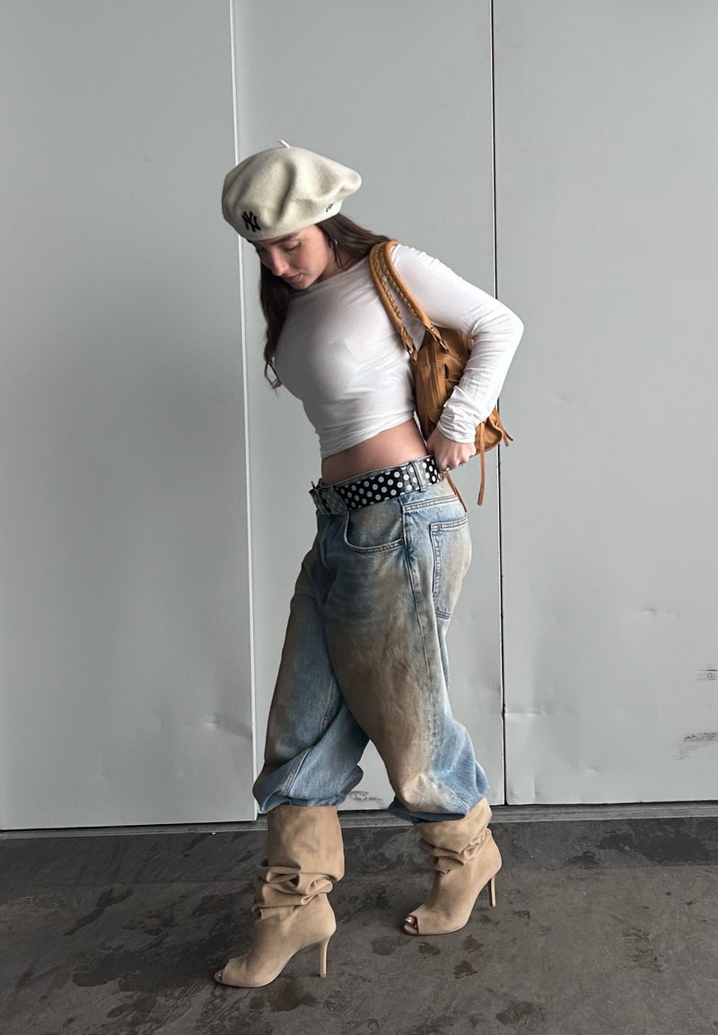 Haut blanc à manches longues, jean bleu délavé ample, ceinture noire à pois, bottes beiges décontractées à talons hauts et béret crème. Sac à main marron.
