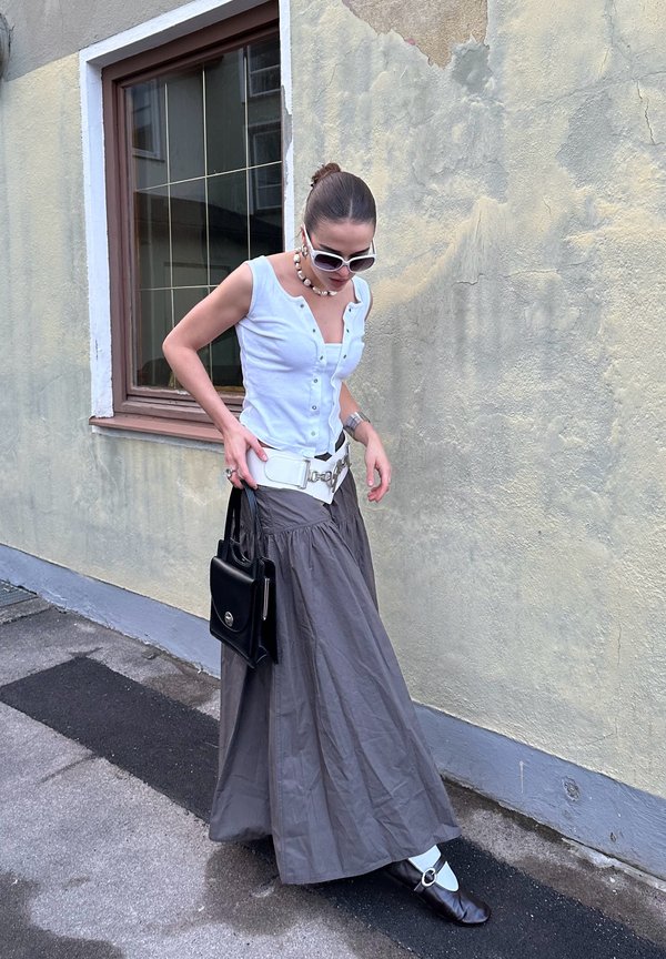 Ανοιχτό μπλε πουκάμισο με κουμπιά, γκρι μακρύ φούστα, λευκή ζώνη, μαύρη τσάντα. Τα αξεσουάρ περιλαμβάνουν γυαλιά ηλίου και ένα κολιέ με χάντρες. Αστική τοποθεσία.